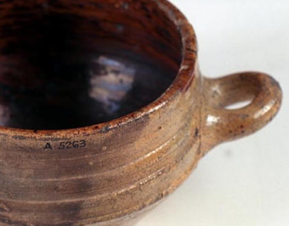 Photograph of a simple high-sided pottery bowl with a brown glaze inside and a single loop handle