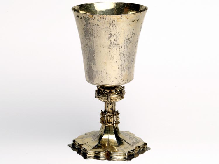 Photograph of a silver chalice, shaped like a wine glass. The top is plain with a wide rim. The stem and base are six-sided with decorative patterns and mouldings.