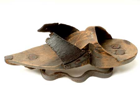 Woman or girl's patten with wooden sole and two black leather straps with holes for ties across the feet. This patten has an undulating curved metal ring under the sole to raise the patten above ground. The toe is pointed and reinforced with metal from the patten ring. This patten has no heel but has a heel socket which measures 2.8 cm. It has a straight sole so it could be worn outdoors. The leather straps are nailed on with three nails and are reinforced. There is a wavy iron ring nailed on at the forepart and two nails at the heel.

