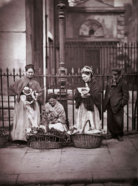 Covent Garden flower women, c. 1877. This group of flower sellers are standing outside St. Paul's Church, their regular spot through generations. Whilst Covent Garden Flower, Fruit and Vegetable Market was a wholesale market, there was room for a few sellers of nosegays to passers-by. Poignantly, Adolphe Smith contrasts the life of these Covent Garden flower sellers with that of Isabelle, the favourite flower-girl of the Paris Jockey Club, who was well rewarded for her services. From a series of 37 photographs published in the book, 'Street Life in London' (1877), with text written by Thomson and the journalist Adolphe Smith. [p.8]

