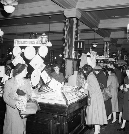 Interior of Selfridges, next to embroidery sets for sale. Women customers sorting through the goods. Christmas 1953
