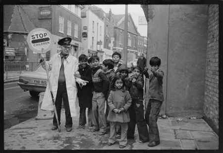 A male crossing patrol warden stands by a group of Asian children outside Christchurch Church of England School, in Spitalfields, 1979.