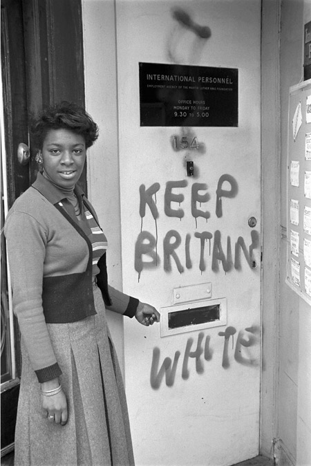 A woman stands outside the door of the Employment Agency of the Martin Luther King Jr. Foundation which has been vandalised and marked 'KEEP BRITAIN WHITE'. As she gestures towards the graffiti, the woman gazes out towards the photographer.