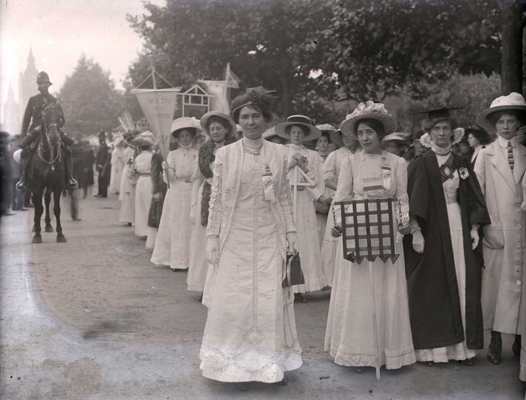Suffragette prisoners prepare to take part in a procession In support of the Conciliation Bill, 23rd July 1910. In the foreground are the leading Suffragettes Emmeline Pethick-Lawrence on the left and Sylvia Pankhurst holding a model of prison gates. To the right in her graduation robes is Emily Wilding Davison. This procession was organised by the Women's Social and Political Union to take place on the anniversary of the day in 1867 when men demonstrating for their inclusion in the Reform Bill pulled down the railings in Hyde Park. Two processions, one from the west and one from the east converged on Hyde Park where 150 speakers addressed at crowd of up to 20,000 from 40 platforms.