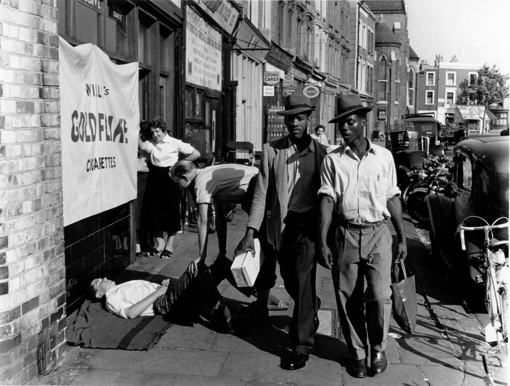 Two black men pass by a man lying on the pavement with another man standing over him and a woman watching, Latimer Road area, North Kensington, 1957.