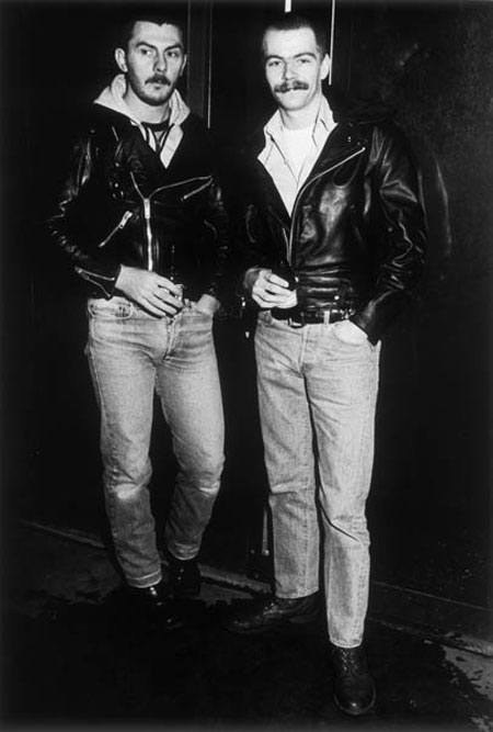 Two young gay men sporting moustaches and black leather jackets posing for the camera outside a pub or club, 1985.
