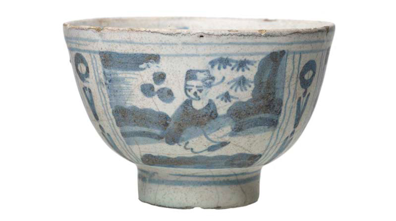 A well-used-looking bowl glazed white with blue patterns on showing a figure and patterns in a Chinese style.