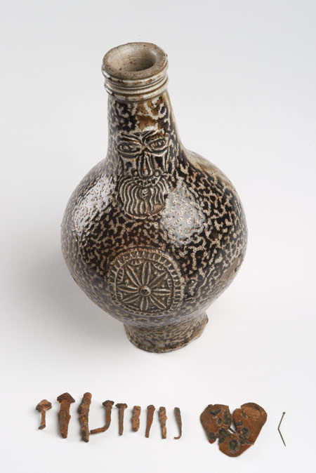 17th century stoneware Bartmann jug (also known as a Bellarmine) made in Frechen, Germany. It has been re-used as a witch bottle. It was found with a heart-shaped piece of felt pierced with pins and eleven nails inside. This would have been a charm to prevent witchcraft. The bottle is decorated with a bearded face mask on the neck and a rosette medallion on the body.

