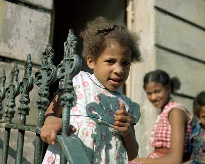 A young girl holding onto railings on a residential street. This photograph is from a series of street scenes by Paul Styles taken in and around the Notting Hill area, often featuring members of the local black community.
