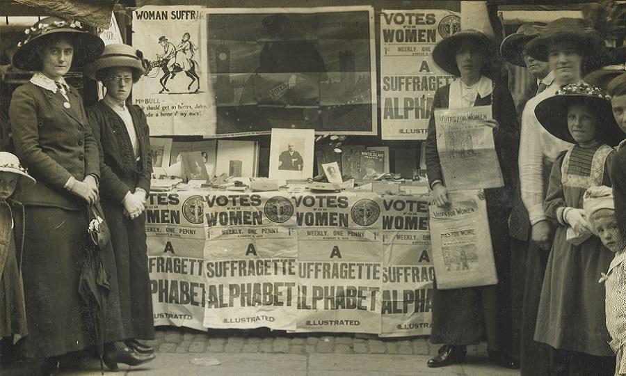 Women and girls stand around a suffragette fundraising stand holding 'Votes for women' posters