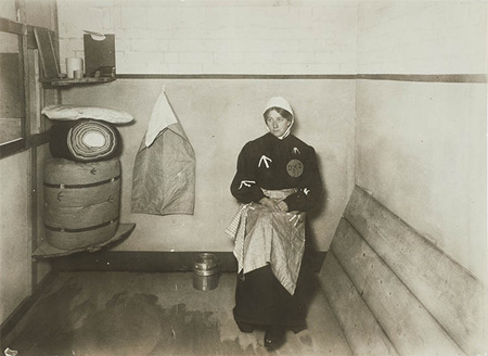 The Suffragette Elsie Howey in a replica prison cell, 1909. Replica prison uniforms were often worn by ex-suffragette prisoners at demonstrations and fund-raising bazaars to highlight the conditions under which imprisoned Suffragettes were held. This image was probably taken at the Women's Exhibition held at Princes Skating Rink in May 1909 where a 'Prison Life' exhibit included a replica prison cell 'peopled' by Suffragettes dressed in replica prison clothing and taking part in prison activities such as sewing.
