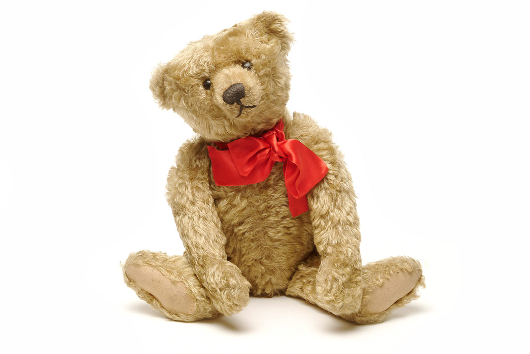 A sandy coloured,jointed teddy bear with button eyes, hump on its back and a Steiff metal button in its ear. There is a red bow around its neck. The donor's eldest son received this teddy bear as a present in 1907, when it was the latest fashionable toy. It makes a growling sound when the stomach is pressed and was made by the Steiff Company in Germany. Sandy coloured plush, probably Excelsior stuffed, with jointed arms and legs, button eyes, hump on back and Steiff metal button in ear. In 1902, Margarete Stieff, a disabled german toy manufacturer, made the first plush toy bears. The name comes from a cartoon of US president Theodore ('Teddy') Roosevelt, who refused to shoot a tethered bear. Teddy bears were an instant hit and have become a traditional childhood toy, beloved by millions.
