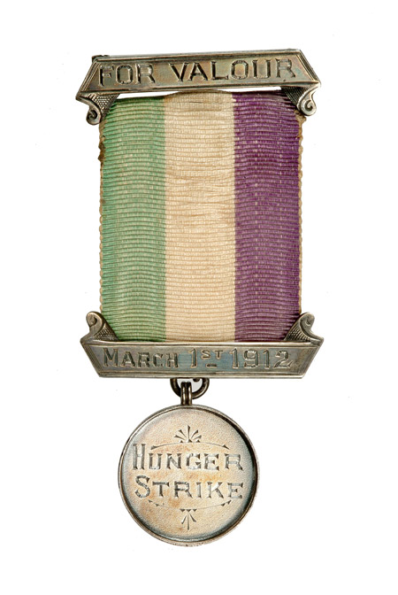 This image shows a suffragette prisoner's silver hunger strike medal with purple, white and green ribbon. It was presented to Emmeline Pankhurst to commemorate her hunger strike when serving a 9 month sentence in Holloway jail for 'conspiracy to incite persons to commit damage to property'.