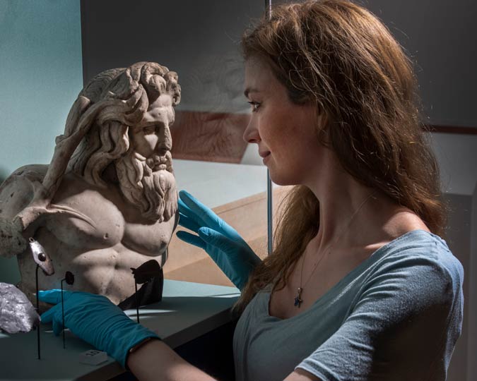 Curator installs a Roman god bust into a glass display case.