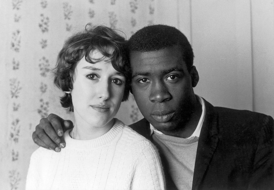 This photograph, entitled 'Notting Hill Couple', is a close-up of a black man and a white woman. He rests his hand on her shoulder while she tilts her head towards his. Both look directly at the camera.