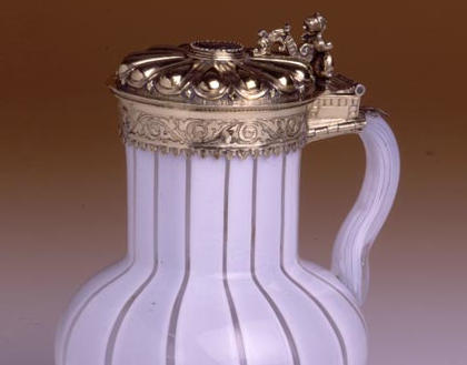 Photograph of a white glass vessel with a round base, narrower top and a looped handle. It has decorative silver bands around the top and bottom, and an elaborately decorated lid silver with a hinge.