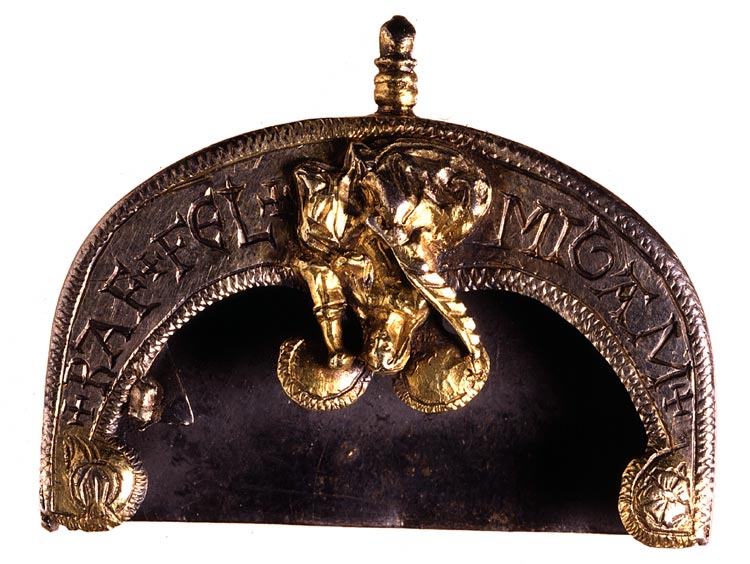Photograph of a D shaped metal object the size of a belt buckle. It has a plain flat base plate with a second flat layer above it following the curved outer edge. This top plate is inscribed, decorated and has a moulded decoration at the centre in the shape of a woman.