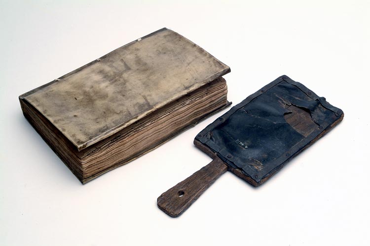 Photograph of a small book, plainly bound in pale leather. To the right is the hornbook; a flat 'paddle' of horn which once displayed sample writing on the front.