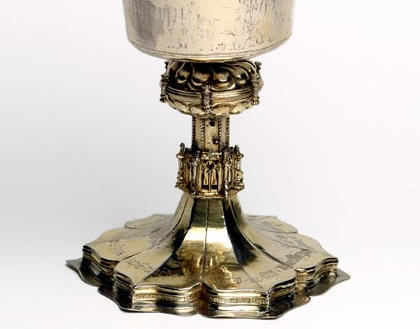 Photograph of a silver chalice, shaped like a wine glass. The top is plain with a wide rim. The stem and base are six-sided with decorative patterns and mouldings