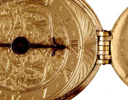 Photograph of the face of an oval, gold-coloured, pocket watch. The numbers are engraved Roman numerals, and have a design of swirling lines and leaves in the centre and around the edge. The hinge and part of the lid can be seen on the right of the picture.