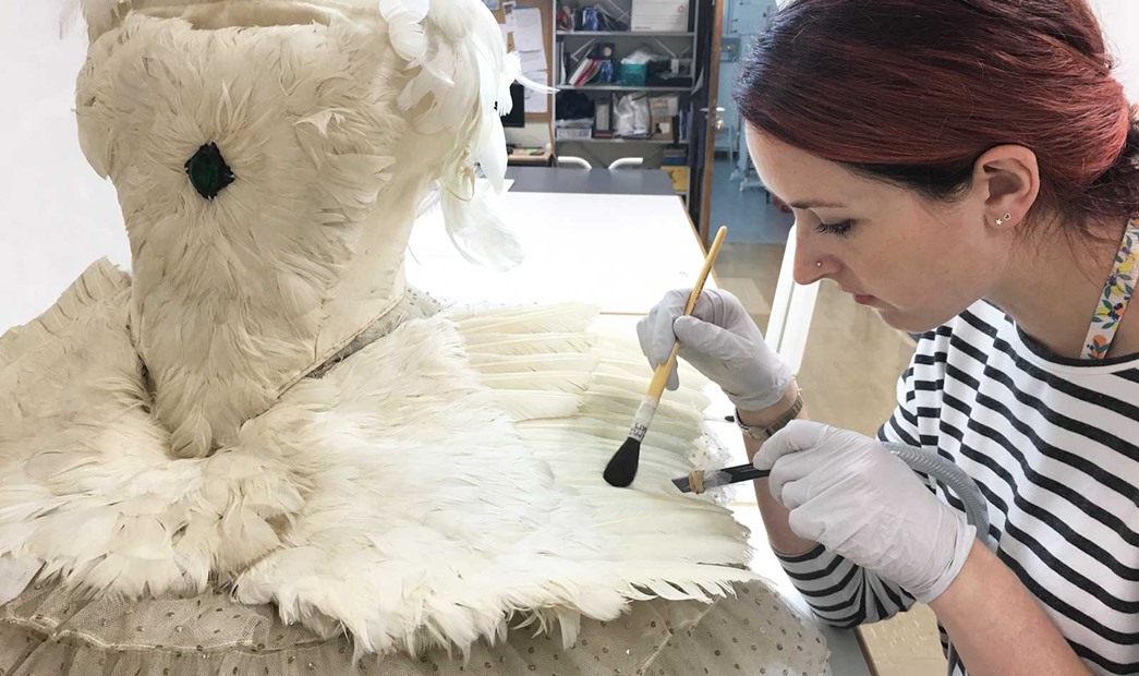 A conservator wearing gloves while cleaning dancer Anna Pavlova's feathered dress, using a low suction vacuum and soft brush.