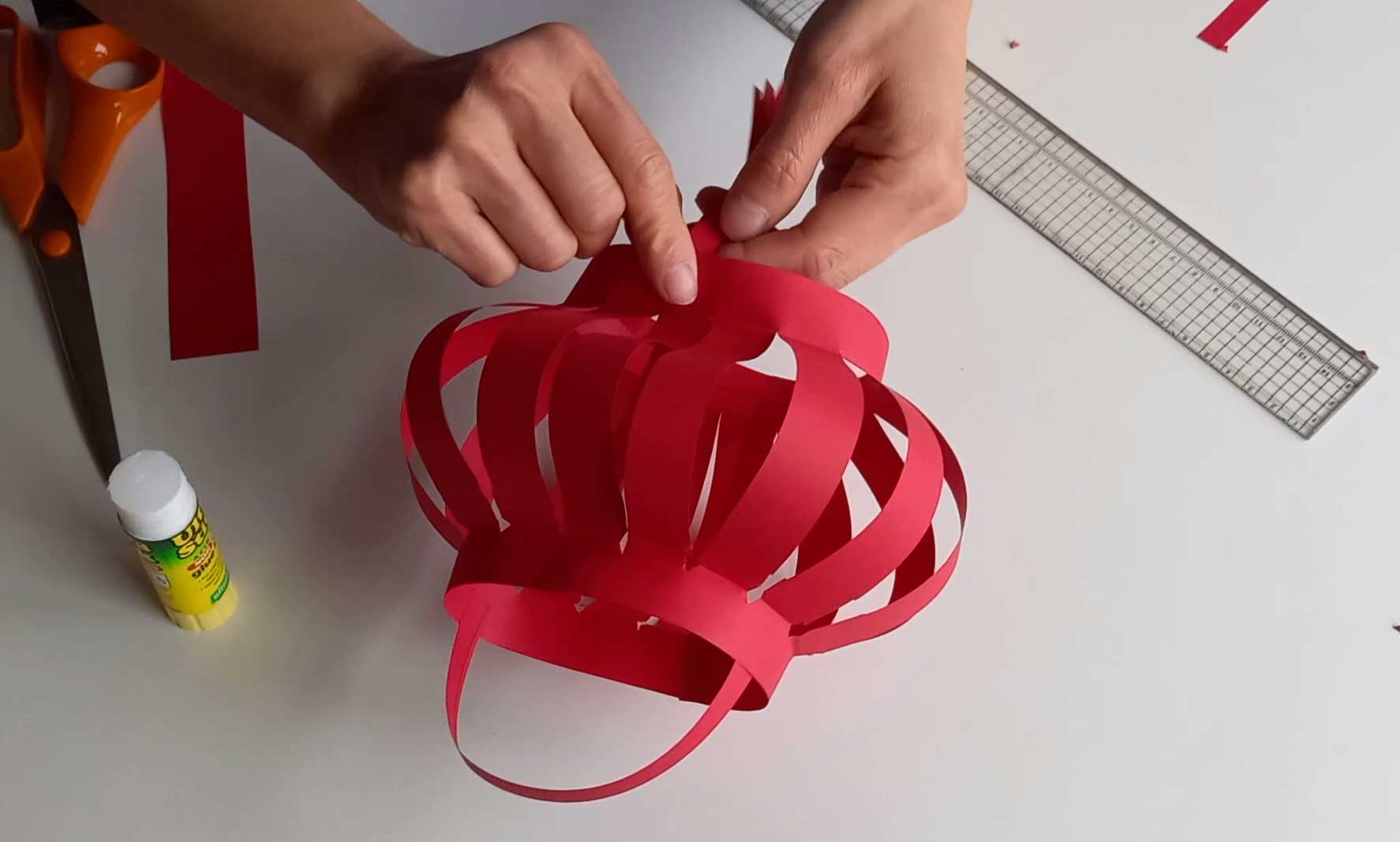 A pair of hands can be seen finishing off making a red cardboard lunar lantern on a table-top next to a glue-stick, ruler and pair of scissors.