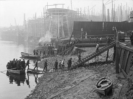 Thames ironworks. Workmen are coming ashore from ferry rowing-boats and crossing the river foreshore via timber causeways and stairs. In the background are ship hulls under construction with timber scaffolding around them.
