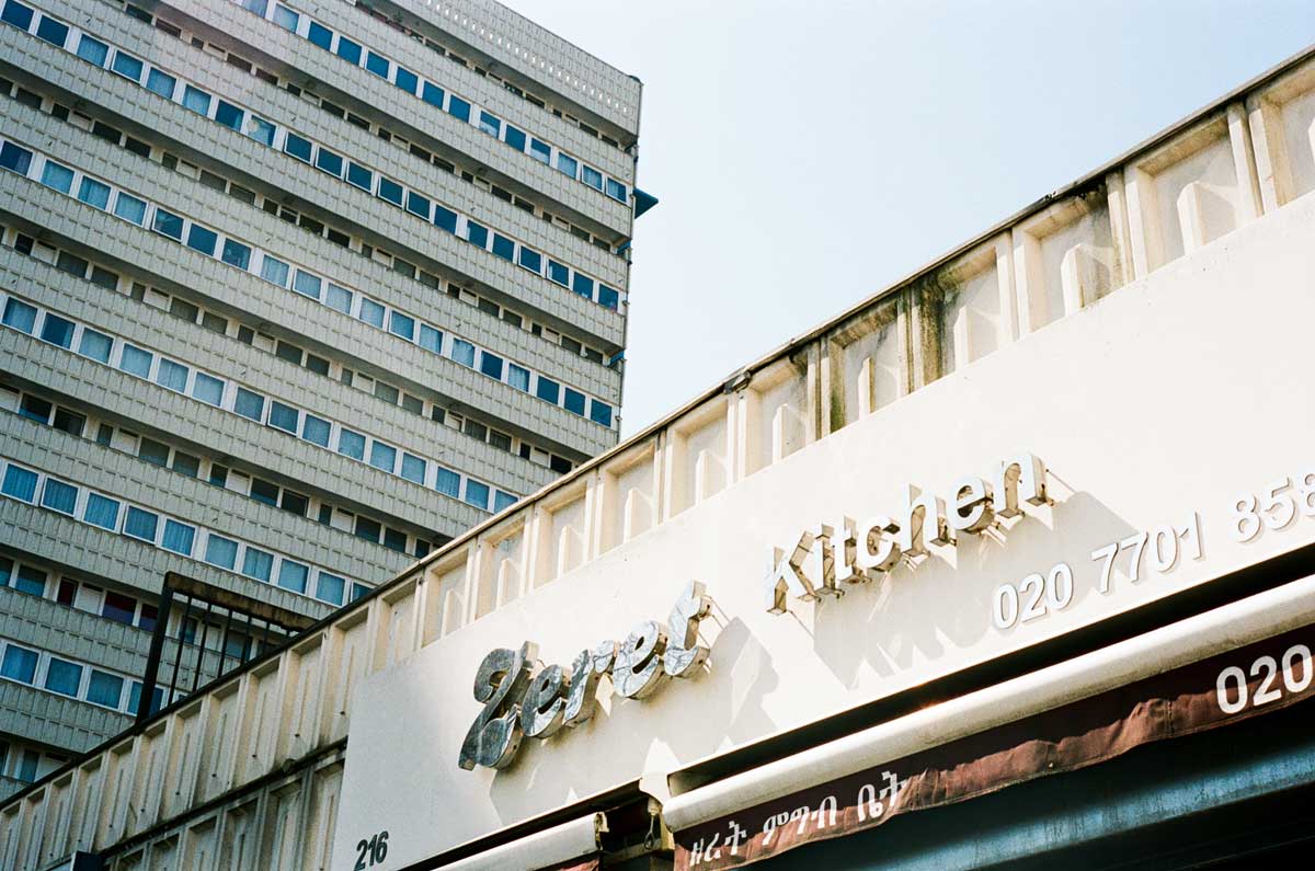 A sign with a phone number on on top of a restaurant, with a block of flats in the background.