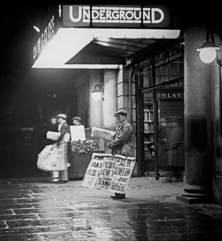 A man selling evening papers outside Charing Cross underground station at night. Mercie Lack.