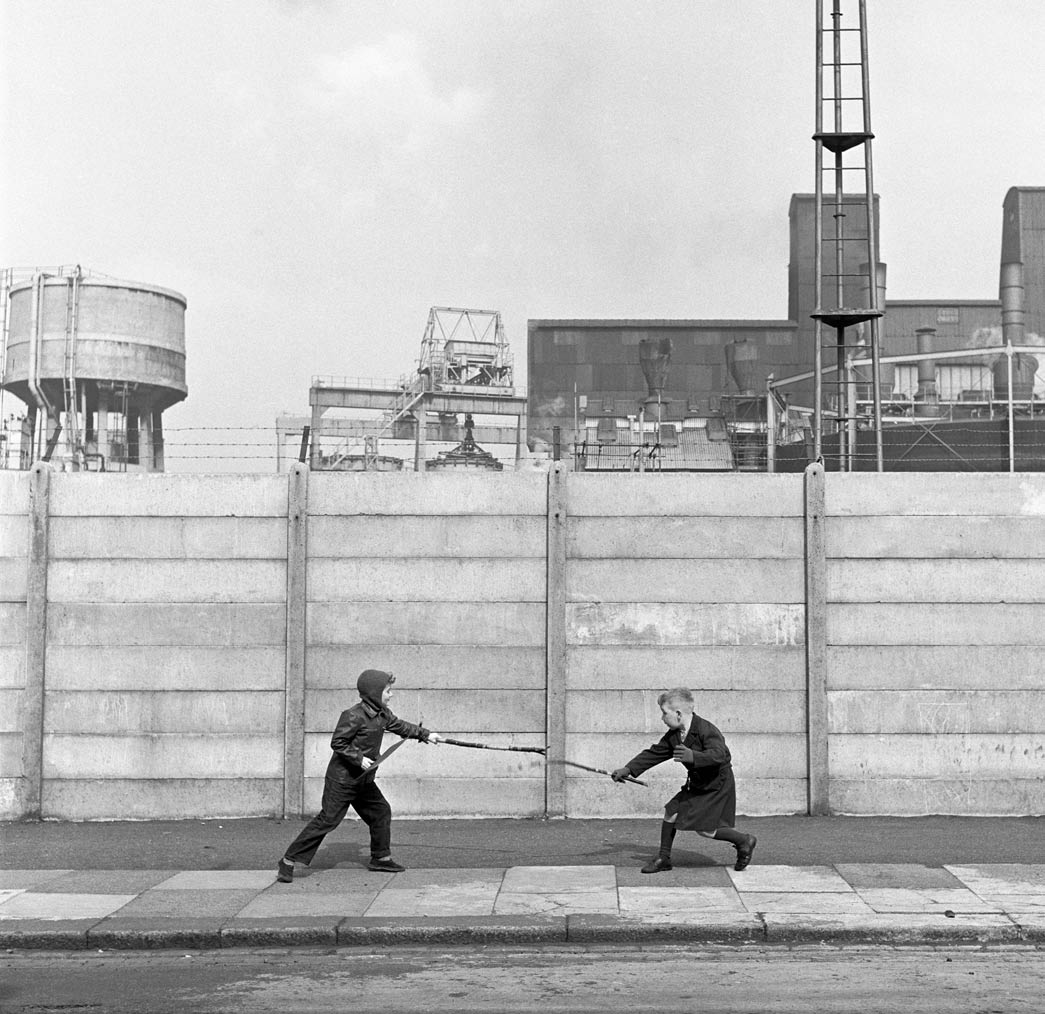 Two boys fighting with sticks in front of a concrete wall. c.1955