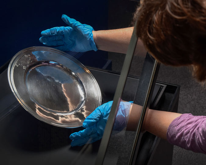 A curator wearing blue gloves installs a silver plate in a glass display case.
