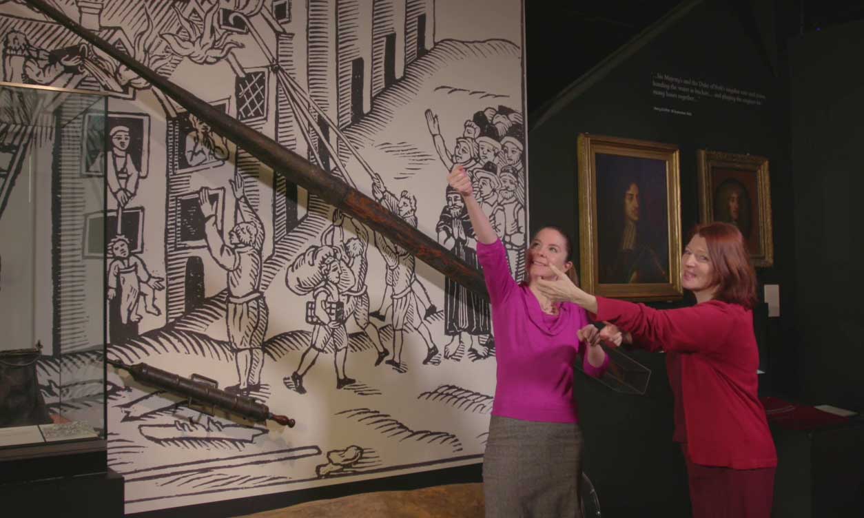 Meriel and Nina pretend to use a fire hook next to a large replica and illustration of the fire.