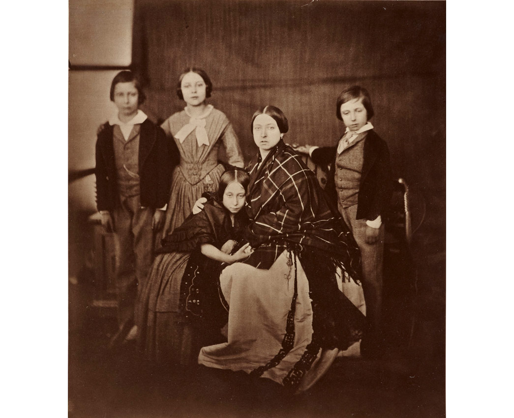 Photograph of Queen Victoria, seated and wearing a checked shawl over her shoulders. She is surrounded by her four eldest children, from left to right, Prince Albert Edward, Prince of Wales, Princess Victoria, The Princess Royal, Princess Alice (seated next to her mother) and Prince Alfred, on the right hand side.
