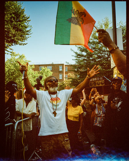 A man wearing Rastafarian clothing waving a Jamaican flag dances at the Notting Hill Carnival.
