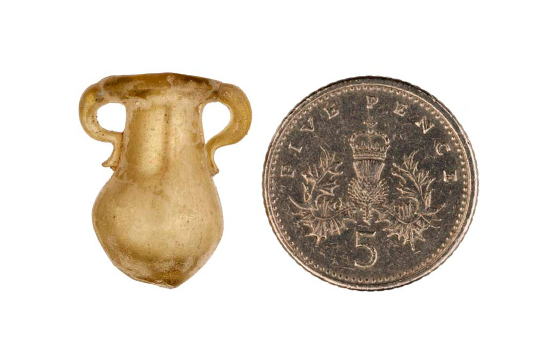 A miniature Roman model of a vase, carved designed to catch tears, photographed next to a 5 pence coin for scale.