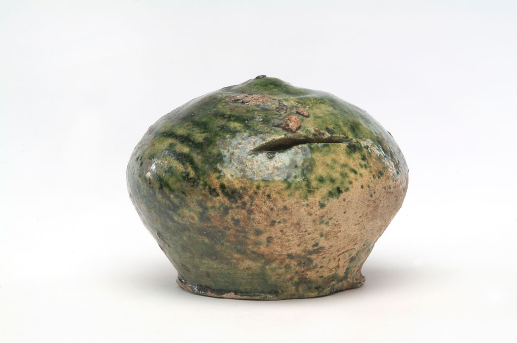 Green glazed ceramic money box. This money box is round and has a slightly pointed top with a flat base. It has an horizontal slot for coins and its body is stained with a green colour. This money box was found complete. It is unusual to find a pottery money box that isn't broken. This type of money box is the medieval equivalent of a piggy bank and normally they are found broken around the slot where people have tried to get the money out. Perhaps this money box was never used. It was made in the Kingston area.
