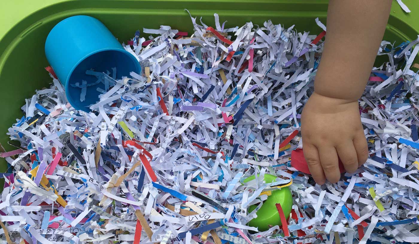 A small hand reaches into a large green plastic tub filled completely with shredded paper, colourful plastic objects and a plastic cup.