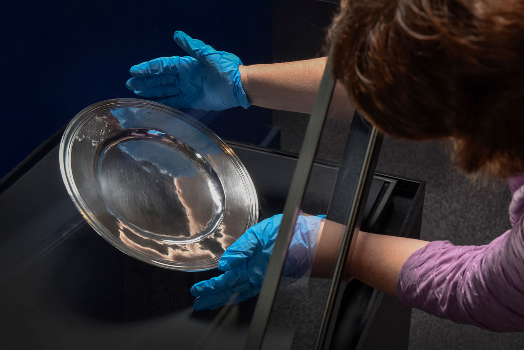 A curator wearing blue gloves installs the Pepys silver plate in a display case.