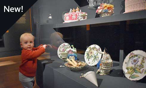 A photograph of a toddler leaning against a museum cabinet containing ceramic plates and ornaments.