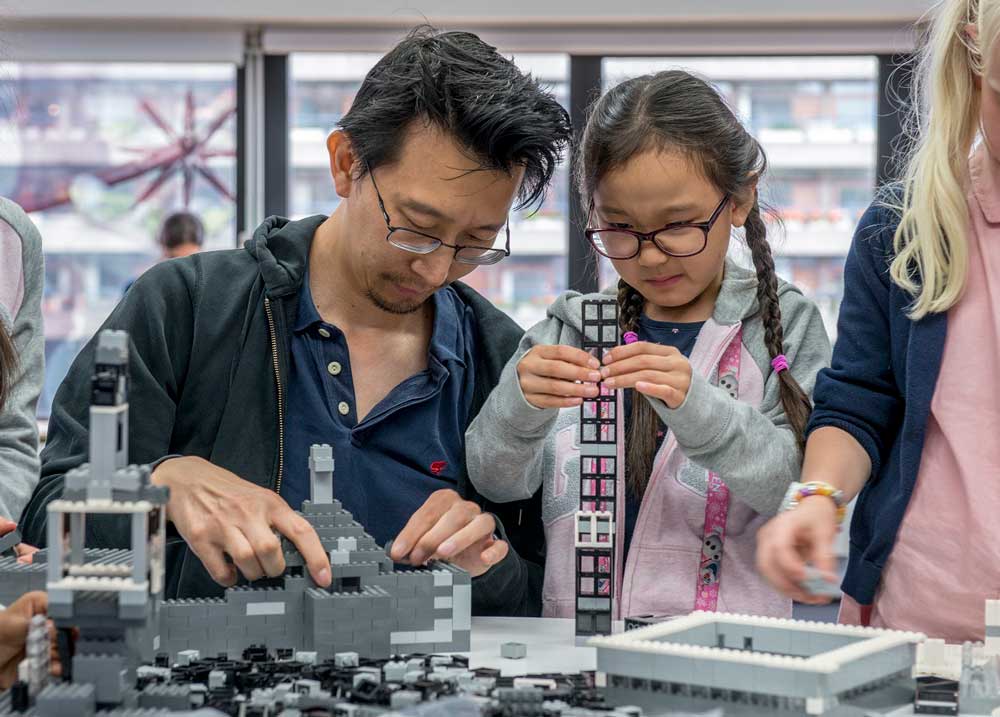 A photograph of an adult and child building architectural structures with grey lego at a museum event.