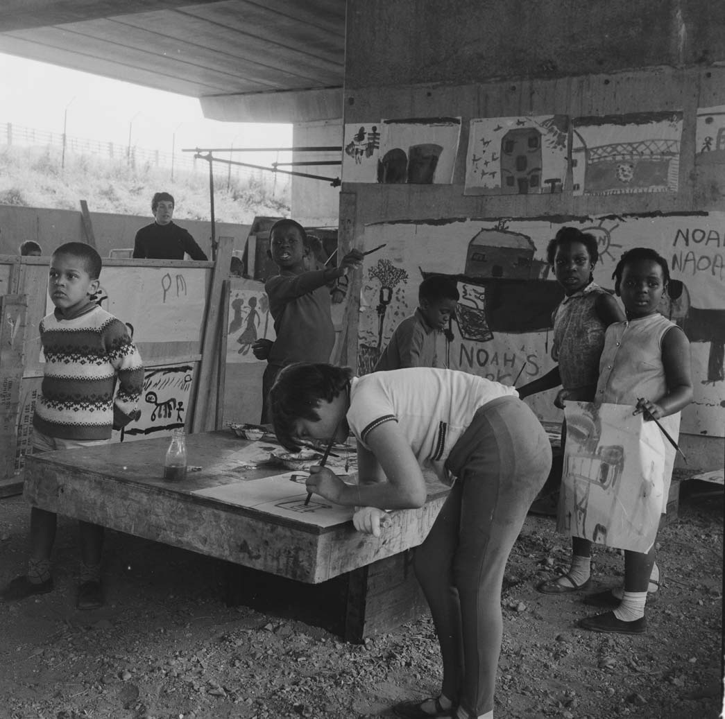 Children using a playspace under the motorway (the Western Avenue extension) in North Kensington in July 1968. Conditions in North Kensington in the 1960s were difficult with poor accommodation, largescale overcrowding and few open spaces. In 1966 the land had been cleared for the building of the motorway and the children and their parents of the London Free School had spontaneously begun to use the space for play. The North Kensington Playspace Group was formed to campaign to make the space permanent. An architect drew up designs for the area and plans were submitted to the Greater London Council. The playspace was opened for the summer holidays at the beginning of August by Sir Laurence Lindo, High Commissioner for Jamaica.