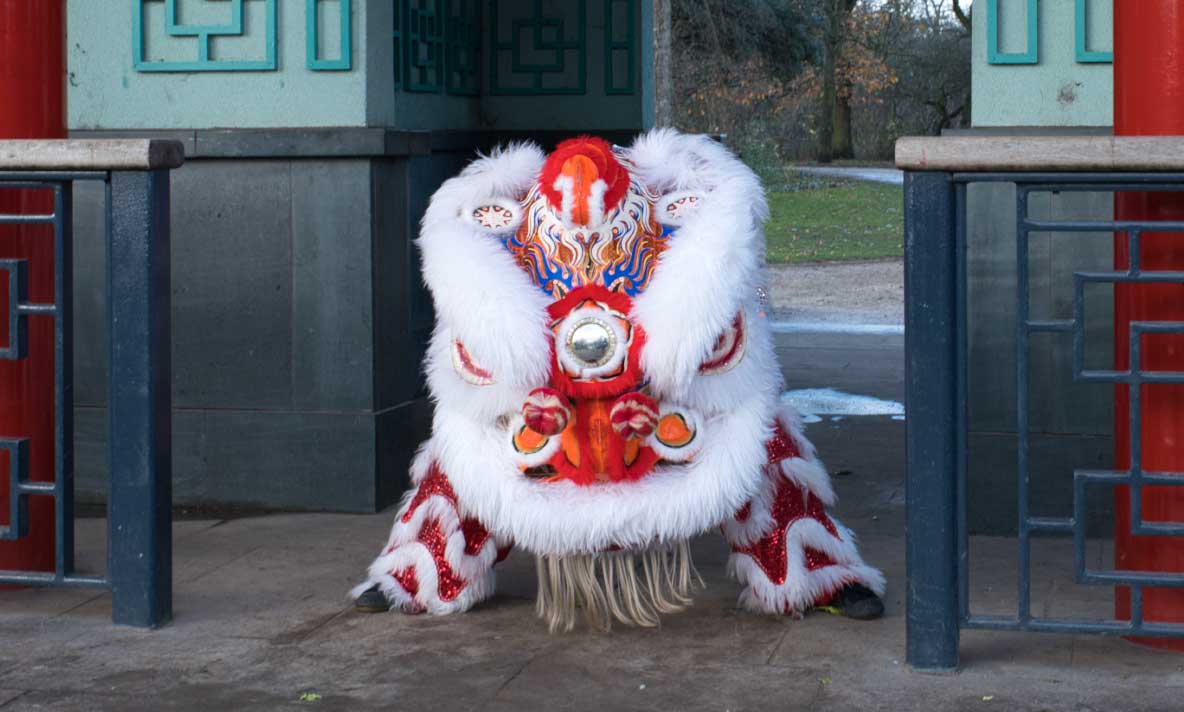 A Lion Dancer in a white and red lion costume lowers their head in between two pillars.