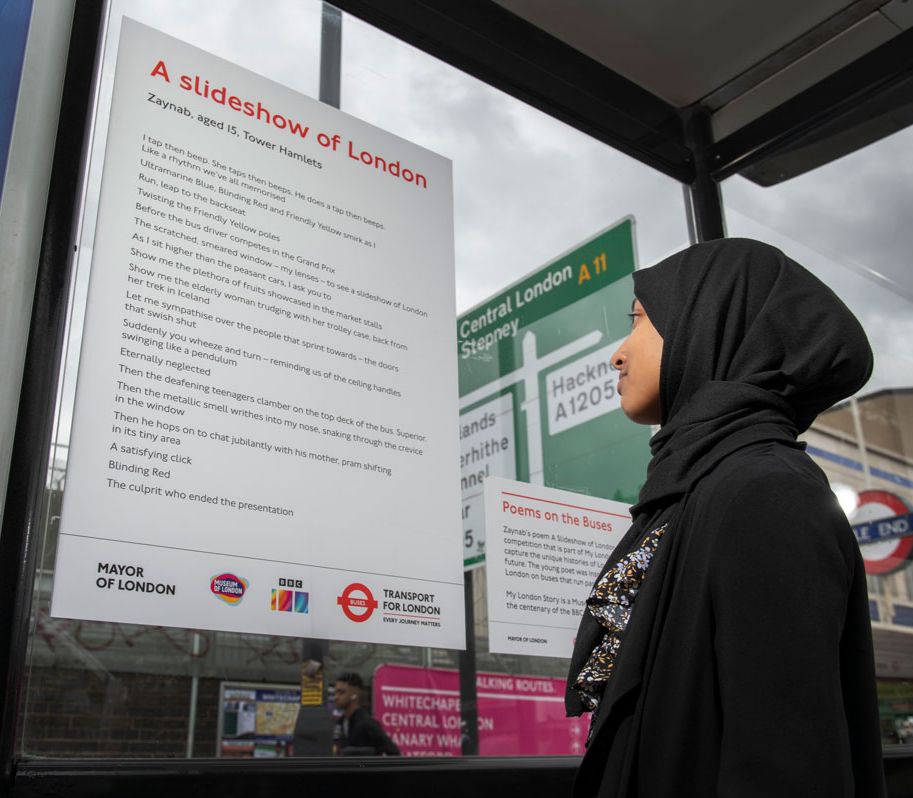 A teenage girl stands looking at her poem on a plaque at a London bus stop.