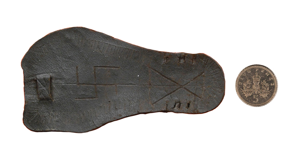 A miniature Roman model of a sandal, carved with a swastika, photographed next to a 5 pence coin for scale.