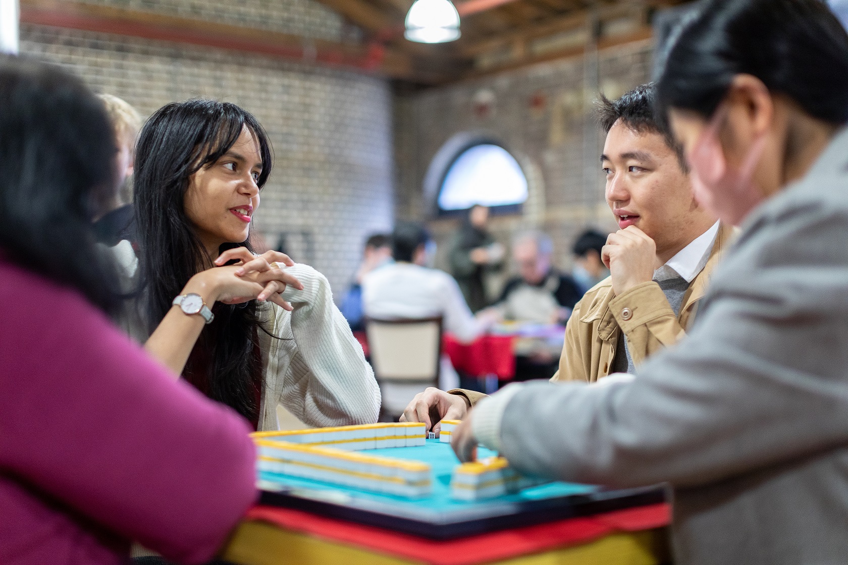 Four people gather around a table and are talking among themselves. One of them is moving one of the pieces of a brightly coloured game on the table.