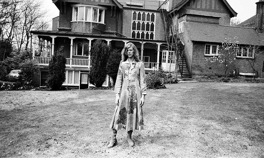 A black and white photo of David Bowie standing in front of a house wearing a dress designed by Mr Fish