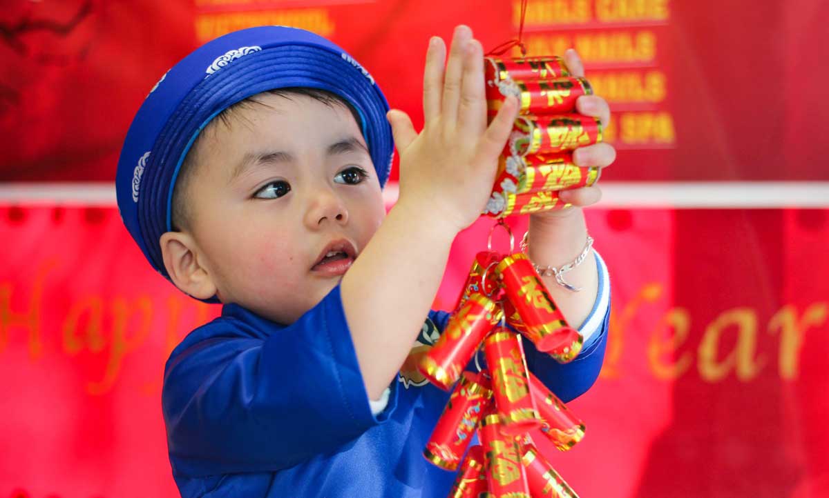 A young child wearing a rich blue robe and hat holds a bright red and gold firecracker up between both hands. Photograph by Anna Nguyen.