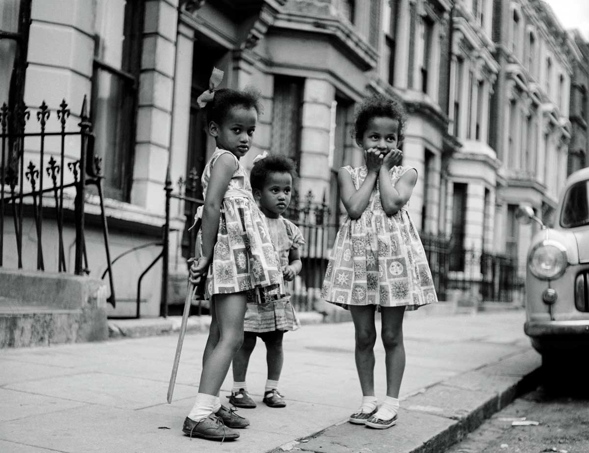 Three young girls, two in matching dresses, play on the pavement in Notting Hill.